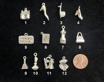 May include: A collection of silver-toned charms, including a castle, high heels, a llama, a dress form, a purse, and a compact. A penny is included for size comparison. The charm labeled "PRINCESS" is also visible.