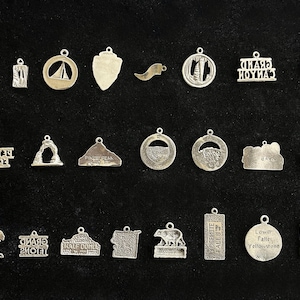 May include: A collection of 25 silver charm pendants featuring various national park and landmark designs. The charms are all silver and have a variety of shapes and sizes. Some of the charms feature text, such as "Grand Canyon" and "Yosemite Falls".