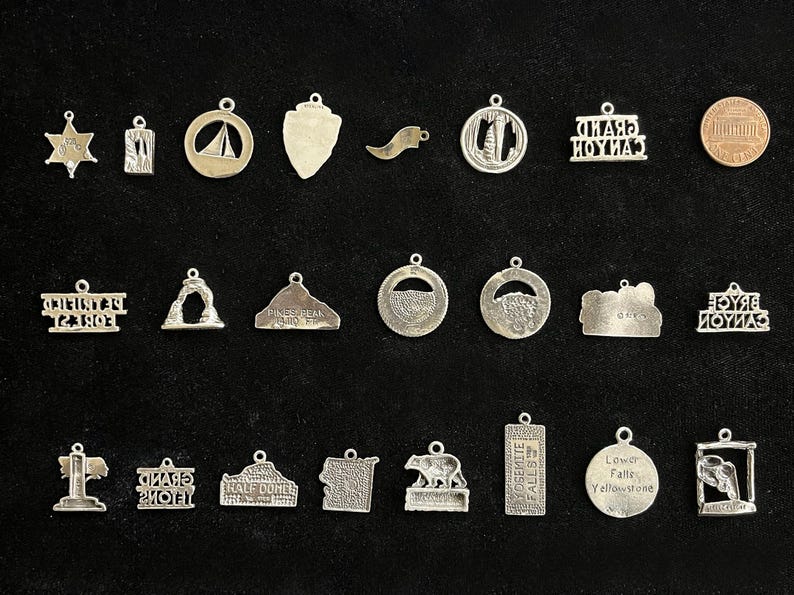 May include: A collection of 25 silver charm pendants featuring various national park and landmark designs. The charms are all silver and have a variety of shapes and sizes. Some of the charms feature text, such as "Grand Canyon" and "Yosemite Falls".