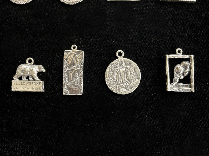 May include: Five silver charms featuring different designs. The charms are rectangular, round, and square. One charm features a bear and the text "Yellowstone National Park".