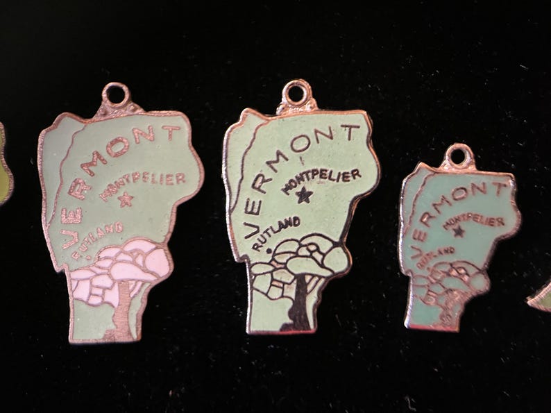 Puede incluir: Tres abalorios de metal verde y rosa con forma del estado de Vermont. Cada abalorio tiene la palabra "Vermont" y los nombres de las ciudades "Montpelier" y "Rutland" escritos en ellos.