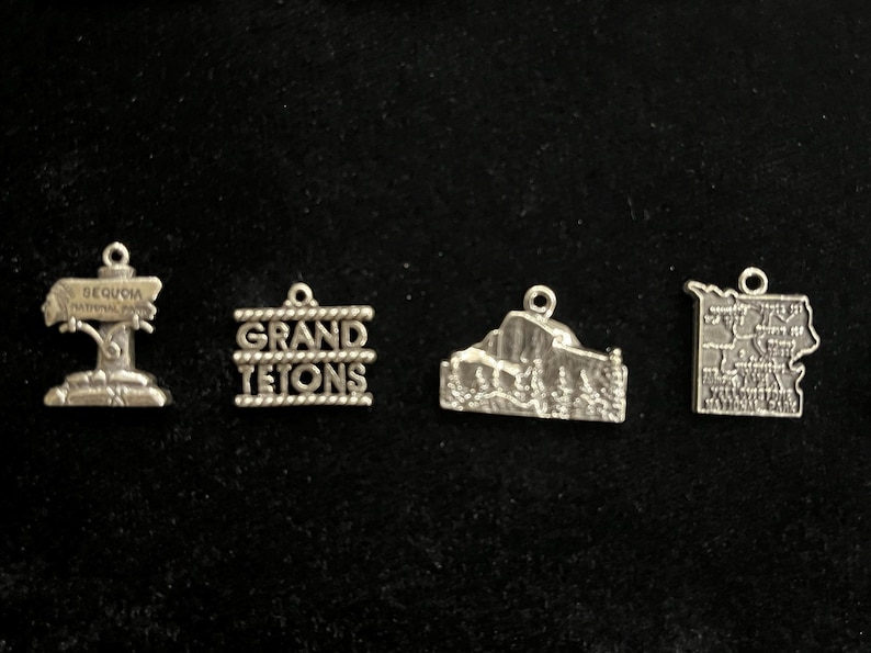 May include: Five silver charms featuring different national parks. The charms are in the shape of signs, mountains, and maps. The charms read "Sequoia National Park", "Grand Tetons", "Yosemite National Park", and "Yellowstone National Park".