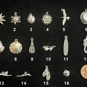 May include: A collection of silver-toned charms with beach and nautical themes. Designs include sun faces, a seashell, a seahorse, a seagull, and a swimmer. A penny is included for size reference.