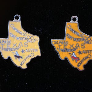 Puede incluir: Dos colgantes de plata con forma del estado de Texas. Los colgantes son amarillos con letras negras y un dise&ntilde;o rojo y blanco. Los colgantes dicen "Texas" y "Friendship" en ellos. Los colgantes est&aacute;n sobre un fondo negro.