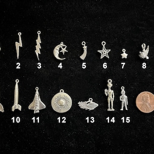 May include: A collection of silver-toned charms with various designs, including a question mark, lightning bolts, a crescent moon and star, a star, a rocket, a spaceship, an alien, and a UFO. A penny is included for size reference.
