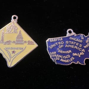 Puede incluir: Dos colgantes vintage con forma de amuleto, uno que representa un contorno amarillo de Washington D.C. con el texto "WASHINGTON" y el otro que representa un contorno azul de los Estados Unidos con el texto "UNITED STATES OF AMERICA" y nombres de ciudades.