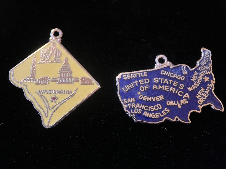 Puede incluir: Dos colgantes vintage con forma de amuleto, uno que representa un contorno amarillo de Washington D.C. con el texto "WASHINGTON" y el otro que representa un contorno azul de los Estados Unidos con el texto "UNITED STATES OF AMERICA" y nombres de ciudades.