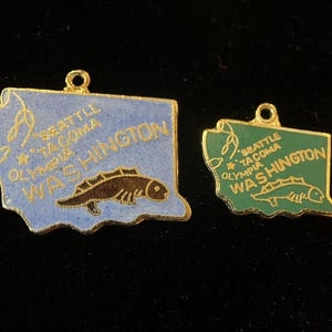 Puede incluir: Dos abalorios de color dorado con forma del estado de Washington, con esmalte azul y verde. Cada abalorio tiene un pez y el texto "Seattle - Tacoma Olympia Washington".