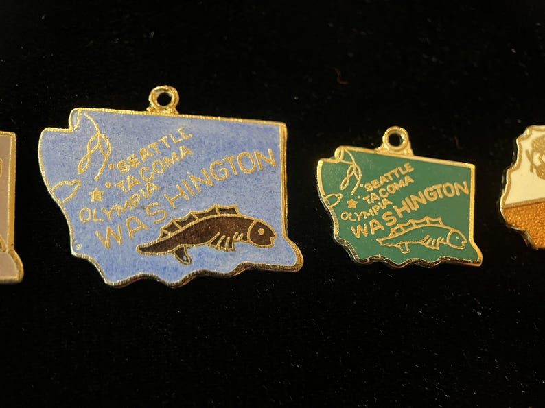 Puede incluir: Dos abalorios de color dorado con forma del estado de Washington, con esmalte azul y verde. Cada abalorio tiene un pez y el texto "Seattle - Tacoma Olympia Washington".