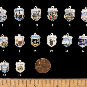May include: A collection of 14 enamel and silver charms shaped like shields, each featuring a different city or state. The charms are numbered 1 through 14. The charms are arranged in three rows, with a ruler and a penny coin below the charms.