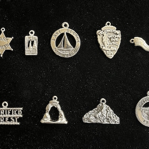May include: A collection of ten silver charms featuring various national park and nature-themed designs. The charms include a star-shaped charm with the words "Park Ranger", a charm with a mountain scene, a charm with a sailboat on a lake, a charm with a mountain range, a charm with a petrified forest, a charm with a mountain peak, a charm with a lake scene, a charm with a mountain arch, a charm with a mountain range, and a charm with a mountain scene.