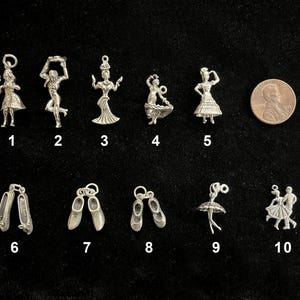 May include: A collection of silver-toned charms with a dance theme. The charms include ballerina figures, ballet shoes, and dancers in various poses. A US penny is included for size reference. The charms are displayed on a black background.