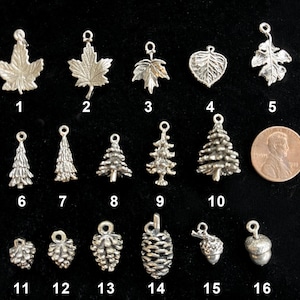May include: A collection of silver-toned charms in various nature-inspired designs, including leaves, pine trees, pine cones, and acorns. A penny is included for size comparison. The charms are numbered 1 through 16.