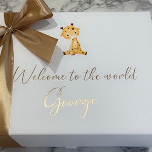 May include: White gift box with a gold ribbon and bow. The box has a gold giraffe illustration and the text "Welcome to the world George" printed on the front.