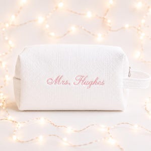 May include: A white rectangular cosmetic bag with a textured fabric finish. The front features the embroidered text "Mrs. Hughes" in a light pink cursive script. The bag includes a side handle and a zip closure on the right side.