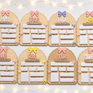 May include: Eight arched wooden signs for school milestones, each featuring a name, a bow at the top, and smaller bows on the sides. The signs include white rectangular spaces for writing details like date, age, height, favorite things, teacher, and future career goals.