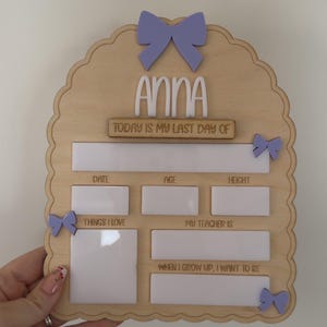 May include: A light-colored wooden arch-shaped sign featuring the name Anna in white lettering. The board includes the text Today is my last day of, Date, Age, Height, Things I love, My teacher is, and When I grow up, I want to be, with white panels.