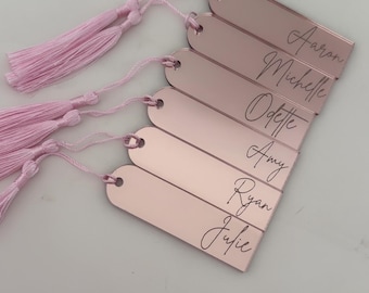 May include: Rose gold rectangular tags with names engraved in a script font. Each tag has a pink tassel attached. Names include Aaron, Michelle, Odette, Amy, Ryan, and Julie.