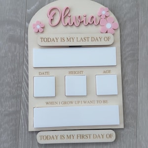 May include: A wooden milestone board with the name "Olivia" in pink script, adorned with pink flower accents. It features sections for "Today is my last day of," date, height, age, "When I grow up I want to be," and "Today is my first day of."