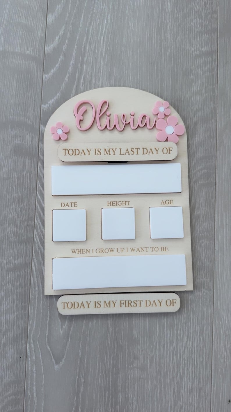 May include: A wooden milestone board with the name "Olivia" in pink script, adorned with pink flower accents. It features sections for "Today is my last day of," date, height, age, "When I grow up I want to be," and "Today is my first day of."