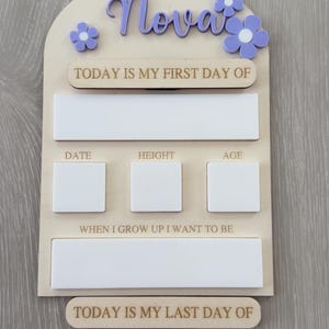 May include: Wooden milestone board with the name "Nova" in purple script and matching flower accents. Includes spaces to record "Today is my first day of", date, height, age, and "When I grow up I want to be". The bottom reads "Today is my last day of".