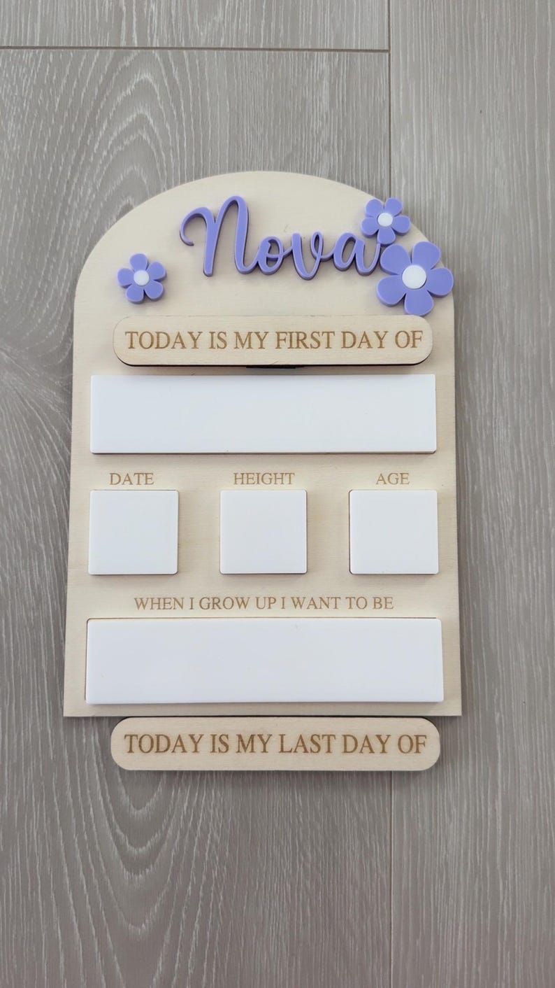 May include: Wooden milestone board with the name "Nova" in purple script and matching flower accents. Includes spaces to record "Today is my first day of", date, height, age, and "When I grow up I want to be". The bottom reads "Today is my last day of".