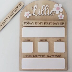 May include: Wooden milestone board with the name "Lillie" and flower accents. Includes spaces for "Date", "Height", and "Age", plus a section for "When I grow up I want to be". A separate wooden sign reads "Today is my last day of".