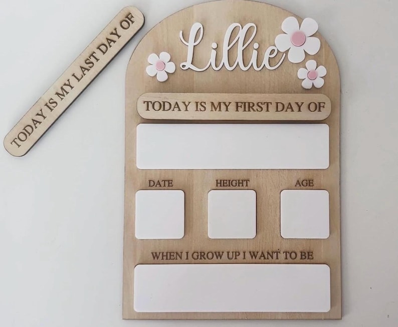 May include: Wooden milestone board with the name "Lillie" and flower accents. Includes spaces for "Date", "Height", and "Age", plus a section for "When I grow up I want to be". A separate wooden sign reads "Today is my last day of".