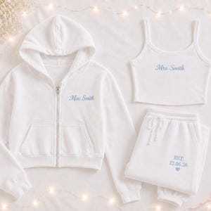May include: A white zip-up hoodie, a white vest top, and white joggers. The hoodie and vest top feature the embroidered text "Mrs. Smith" in light blue. The joggers feature the embroidered text "EST. 22.06.26" and a small blue heart.