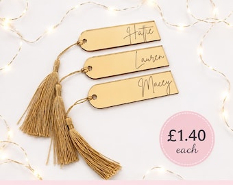May include: Three rectangular gold-toned mirrored bookmarks with rounded ends, each featuring a gold tassel attached through a circular hole. The names Hattie, Lauren, and Macey are engraved in a black script font on the surface of each bookmark.