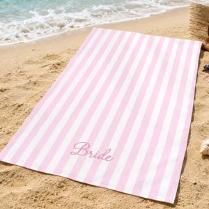 May include: A rectangular beach towel featuring a pattern of vertical light pink and white stripes. The word Bride is embroidered in a cursive script font in a matching pink thread near the bottom center of the towel fabric surface.