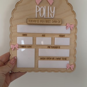 May include: A scalloped wooden sign featuring the name Polly, a large pink bow at the top, and smaller pink bows on the sides. It includes white dry-erase sections for text: Today is my first day of, Date, Age, Height, Things I love, My teacher is, and When I grow up, I want to be.