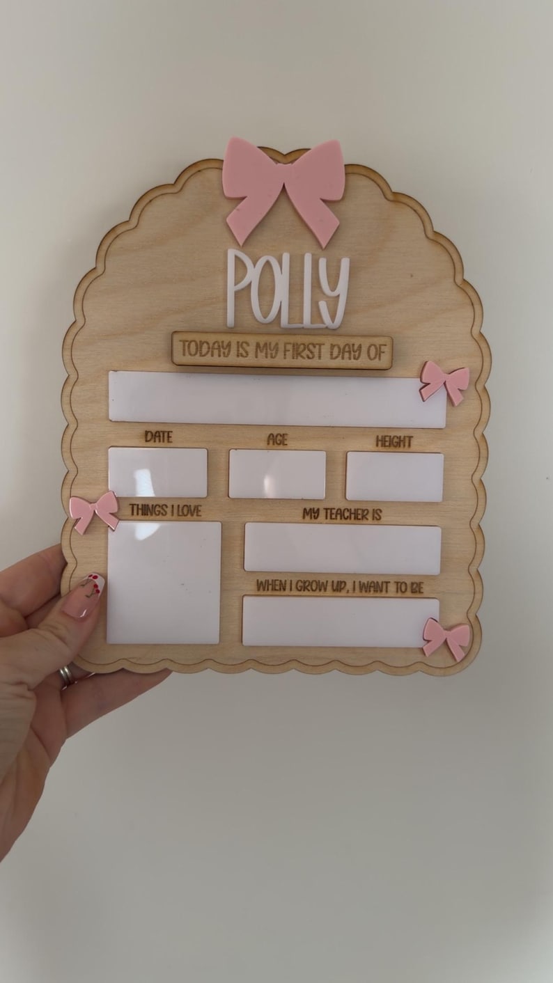 May include: A scalloped wooden sign featuring the name Polly, a large pink bow at the top, and smaller pink bows on the sides. It includes white dry-erase sections for text: Today is my first day of, Date, Age, Height, Things I love, My teacher is, and When I grow up, I want to be.