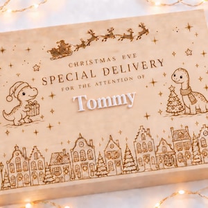 May include: A rectangular light-coloured wooden box featuring laser-engraved illustrations of a dinosaur in a Santa hat, a dinosaur with a scarf, a Christmas tree, and a village scene. The text reads CHRISTMAS EVE SPECIAL DELIVERY FOR THE ATTENTION OF Tommy.