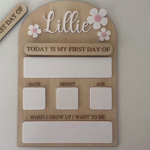 May include: Wooden milestone board with the name "Lillie" and daisy accents. Includes spaces for "Today is my first day of," date, height, age, and "When I grow up I want to be." A separate wooden piece reads "Today is my last day of."