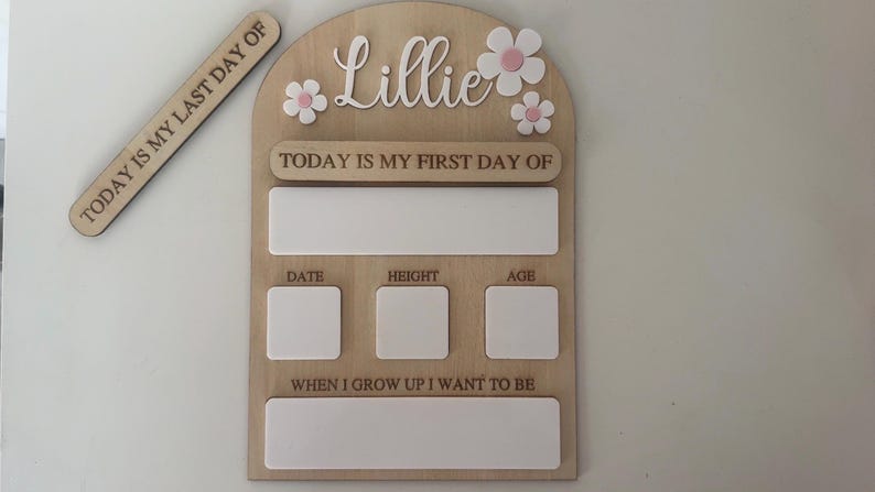 May include: Wooden milestone board with the name "Lillie" and daisy accents. Includes spaces for "Today is my first day of," date, height, age, and "When I grow up I want to be." A separate wooden piece reads "Today is my last day of."