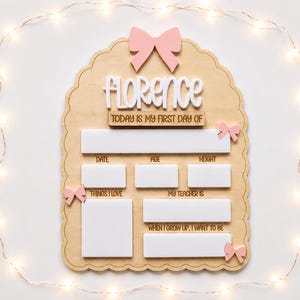 May include: A scalloped wooden milestone board featuring the name Florence in white script, a pink bow at the top, and smaller pink bows on the sides. It includes white rectangular spaces for writing details like date, age, height, and interests.