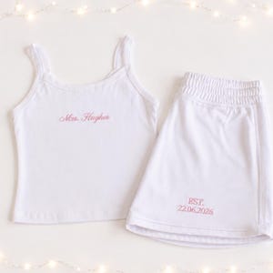 May include: A white vest top and matching shorts set featuring pink embroidered text. The vest top displays the name Mrs. Hughes, while the shorts feature the text EST. 22.06.2026. The garments are made of a plain white fabric with a simple design.