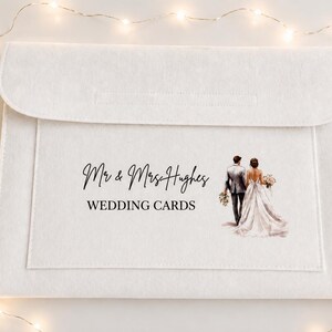May include: A white felt envelope-style card holder featuring the printed text "Mr & Mrs Hughes" and "WEDDING CARDS" in black, accompanied by a color illustration of a couple in wedding attire walking away from the viewer.