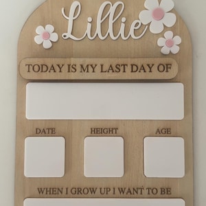 May include: Wooden milestone board with the name "Lillie" in cursive, adorned with white and pink flowers. Includes sections for "Today is my first day of," "Today is my last day of," date, height, age, and "When I grow up I want to be."