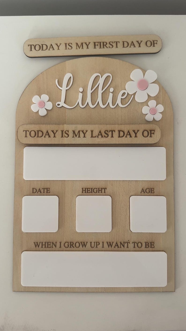 May include: Wooden milestone board with the name "Lillie" in cursive, adorned with white and pink flowers. Includes sections for "Today is my first day of," "Today is my last day of," date, height, age, and "When I grow up I want to be."