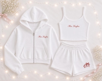 Puede incluir: Una sudadera blanca con cremallera, una camiseta de tirantes blanca y unos pantalones cortos blancos. La sudadera y la camiseta llevan el texto bordado en rojo Mrs. Hughes. El pantalón corto tiene un diseño bordado en rojo de dos corazones con un lazo y las iniciales LH.