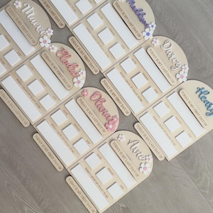 May include: Wooden milestone boards with names like Nova, Maeve, and Elodie, decorated with floral accents. Each board has spaces for date, height, and age, with the phrases "Today is my first day of" and "Today is my last day of".