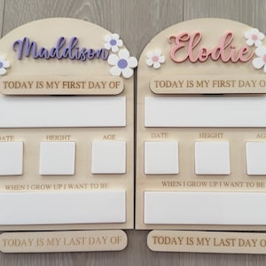 May include: Two wooden first and last day of school signs. One sign has the name "Maddison" in purple script with white and purple flowers. The other sign has the name "Elodie" in pink script with white and pink flowers. Each sign has space for date, height, age, and a statement.