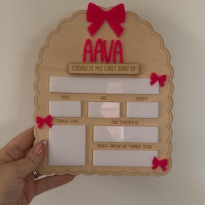 May include: A scalloped wooden sign featuring the name AAVA in pink acrylic, with the text TODAY IS MY LAST DAY OF, DATE, AGE, HEIGHT, THINGS I LOVE, MY TEACHER IS, and WHEN I GROW UP, I WANT TO BE. The sign is decorated with four small pink bows.
