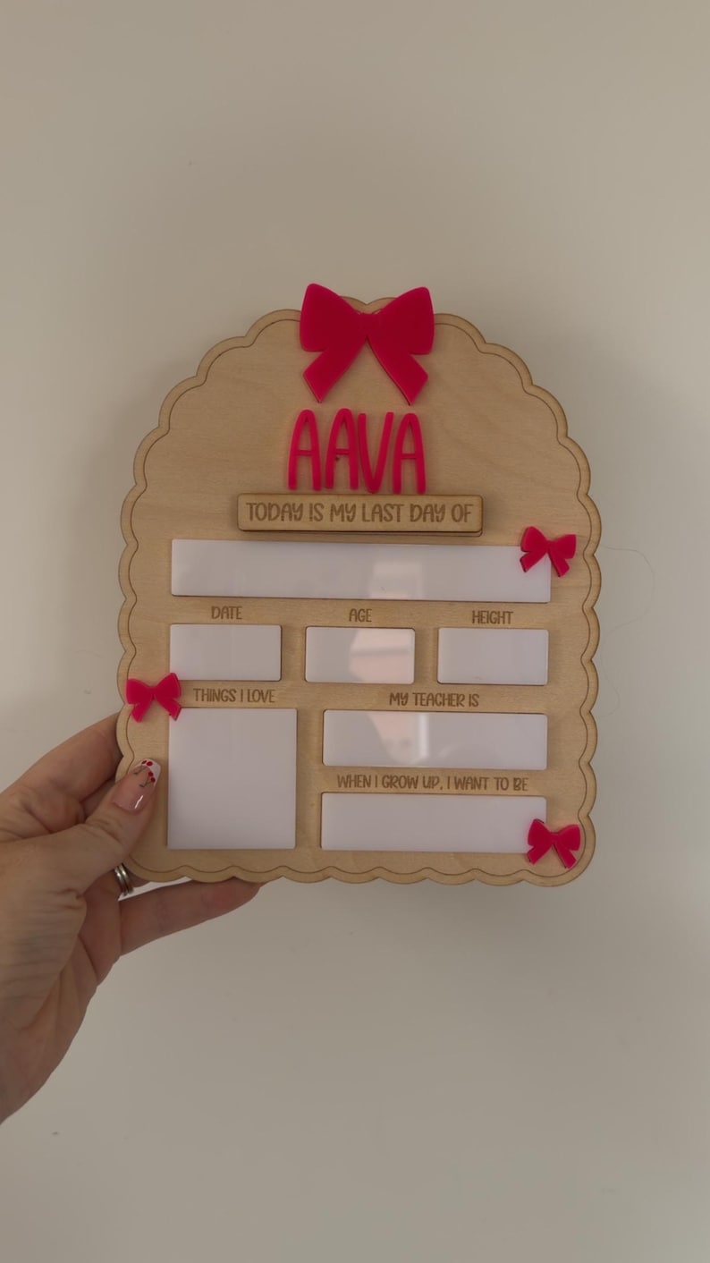 May include: A scalloped wooden sign featuring the name AAVA in pink acrylic, with the text TODAY IS MY LAST DAY OF, DATE, AGE, HEIGHT, THINGS I LOVE, MY TEACHER IS, and WHEN I GROW UP, I WANT TO BE. The sign is decorated with four small pink bows.