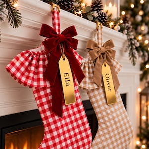 May include: Two Christmas stockings featuring a gingham pattern, one in red and white and the other in tan and white. Each stocking has a ruffled top, a velvet bow, and a gold-toned metal name tag engraved with the names Ella and Oliver.