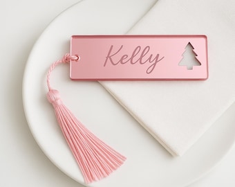 May include: A pink rectangular place card with the name "Kelly" engraved on it, along with a cut-out Christmas tree design. A pink tassel is attached to the card. The card rests on a white plate and napkin.