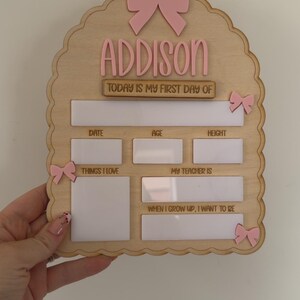 May include: A light wood arch-shaped sign with pink bow accents and the name ADDISON. It features white dry-erase sections for date, age, height, things I love, my teacher is, and when I grow up, I want to be, with the text TODAY IS MY FIRST DAY OF.