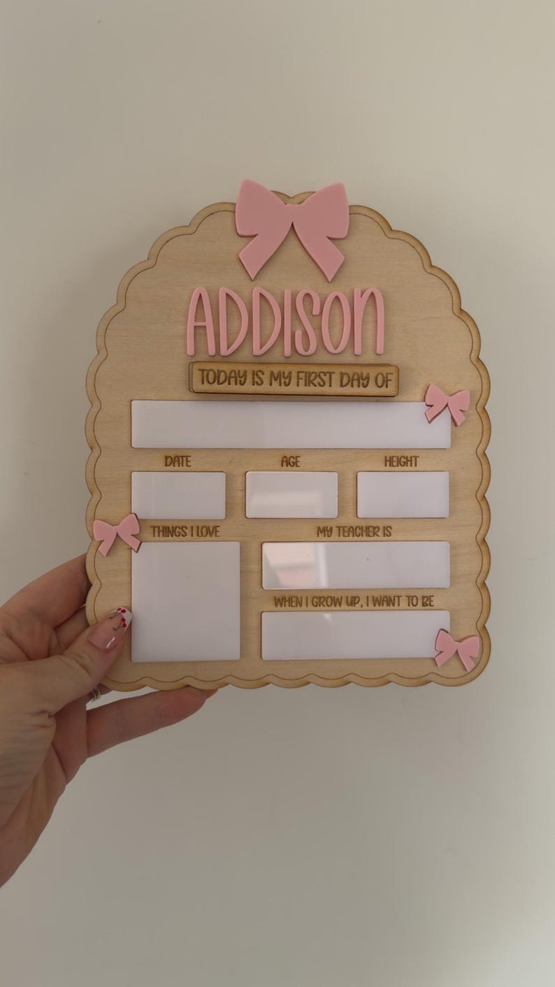 May include: A light wood arch-shaped sign with pink bow accents and the name ADDISON. It features white dry-erase sections for date, age, height, things I love, my teacher is, and when I grow up, I want to be, with the text TODAY IS MY FIRST DAY OF.